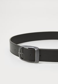 Ceinture en cuir noir présentant une boucle en métal argenté rectangulaire, une texture lisse et plusieurs trous d'ajustement le long de la sangle.