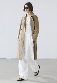 Beige Trenchcoat mit schwarzen Knöpfen, weißem T-Shirt, weißen weiten Hosen und schwarzen Sandalen. Die Accessoires umfassen eine Sonnenbrille und eine Halskette.
