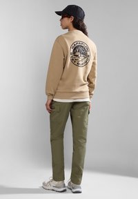 Felpa beige con grande logo scuro sulla schiena, abbinata a pantaloni cargo verde oliva e sneakers beige. Un cappellino nero completa il look.