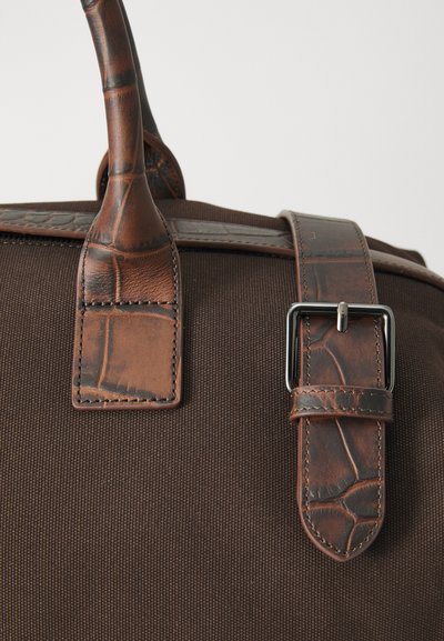 Brown canvas bag featuring a textured brown leather strap and handle, with metal buckle accents for closure.