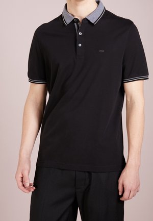 Man draagt een zwart polo shirt met korte mouwen, een grijze kraag en witte biezen, gecombineerd met een zwarte broek, staand tegen een effen achtergrond.