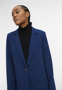Vrouw met gevlochten haar in een blauwe blazer over een zwarte coltrui, iets naar links gericht tegen een effen lichte achtergrond.