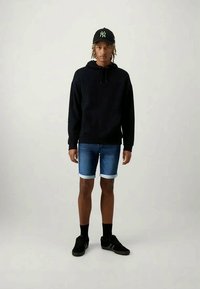 Svart hettegenser, denimshorts med rullekanter, og svarte joggesko. Kapsen er svart med en grønn logo. Avslappet antrekksdesign.