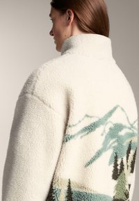 Kremowa kurtka z polaru z wysokim kołnierzem i wzorem krajobrazu górskiego w kolorach zielonym i czarnym na plecach. Miękka, teksturowana powierzchnia.