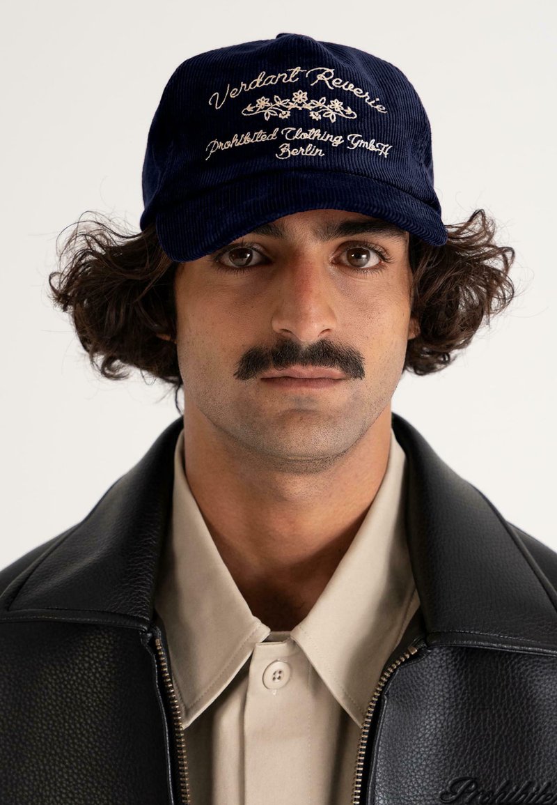Man with curly hair and mustache wearing navy cap embroidered with "Verdant Reverie," beige shirt, and black leather jacket against a neutral background.