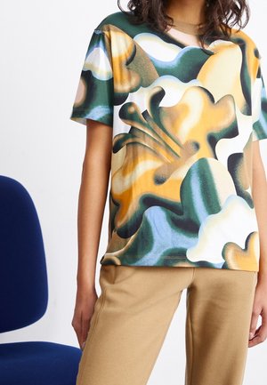 Osoba ubrana w luźną koszulę na krótki rękaw w kwiatowy wzór w odcieniach zieleni, żółci, bieli i brązu, w połączeniu z beżowymi spodniami.