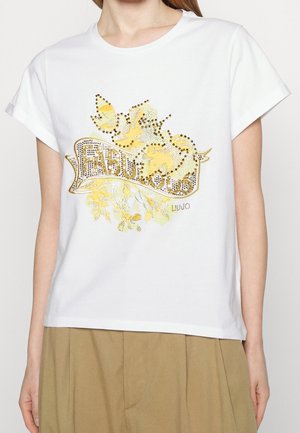 Biała koszulka z żółtym kwiatowym wzorem i złotym napisem z cyrkoni „Fabulous”, noszona z oliwkowymi spodniami.