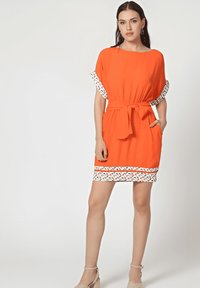Vestido naranja con mangas cortas, cinturón en la cintura y un intrincado borde blanco con patrón en el dobladillo y los bordes de las mangas. Incluye bolsillos frontales.