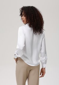 Blusa blanca de mangas largas, textura suave, puños abotonados y dobladillo curvado. Combinada con pantalones beige ajustados.