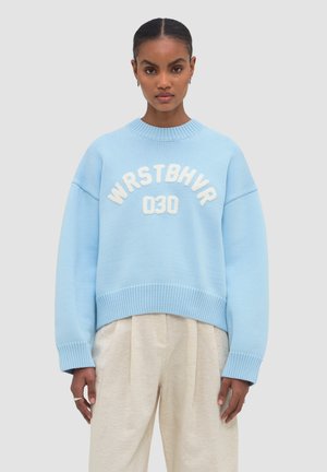 Woman wearing light blue oversized sweater with white "WRSTBHVR 030" text and loose beige pants, standing against gray background.