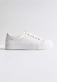 Vit segelsko av kanvas med rund tå, texturerad yta, gyllene öglor och vita snören. Har en platt vit gummisula för bra grepp.