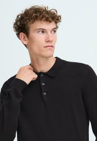 Črna pletena polo majica z dolgimi rokavi, s kragnim izrezom in platinimi gumbi. Gladka tekstura z rebrastimi manšetami.
