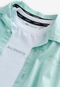 Camicia collare verde menta sovrapposta a una maglietta bianca, con la scritta "ALLSAINTS" stampata in verde chiaro. Tessuto morbido, apertura con bottoni.