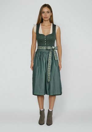 Frau in einem grünen traditionellen Dirndl mit weißer transparenter Bluse und gemustertem Gürtel, stehend vor einem schlichten grauen Hintergrund.