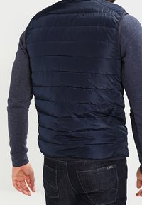 Marinblå pufferväst med quiltat mönster, lätt material och en slät finish. Bärs över en långärmad grå tröja, utan detaljer på ryggen.