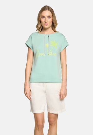 Kobieta ubrana w jasnozielony top z krótkim rękawem z żółtym napisem „Good Vibes” i palmami, w białych szortach do kolan, stojąca prosto.