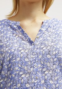 Modrá a bílá květinová košile na knoflíky s výstřihem do V, lehkým materiálem a jemnými vzory listů. Knoflíky jsou malé a kulaté.