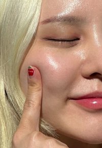 Läheltä kuvattu kasvo, jossa on sileä, hehkuva iho. Vaaleat hiukset ja hienovarainen huuliväri; sormi koskettaa poskea, jossa on punainen mansikkakynsilakka.