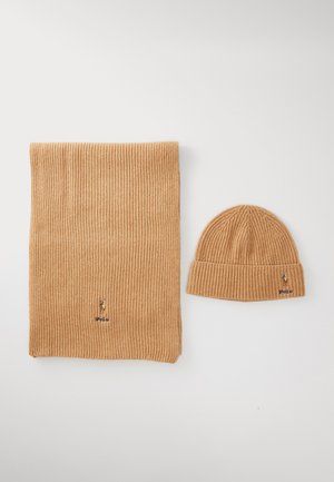 Écharpe en tricot côtelé beige et bonnet assorti avec un petit logo brodé, disposés sur un fond clair.
