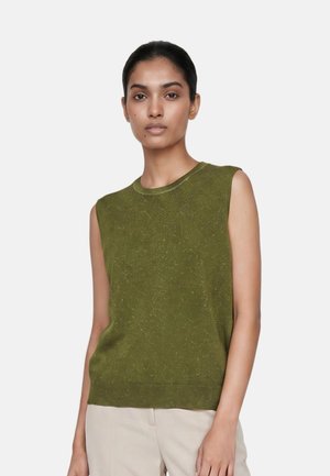 Vrouw met een mouwloos olijfgroen topje en lichtbeige broek, staand tegen een effen witte achtergrond.