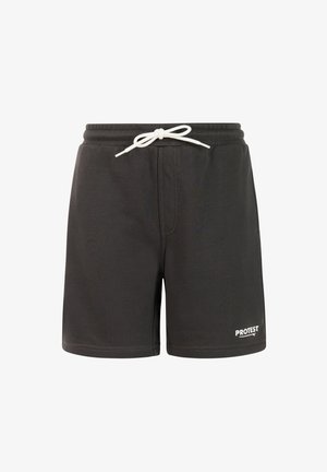 Jonge persoon die staat en een blauw t-shirt, zwarte korte broek, grijze sokken en beige sneakers draagt tegen een effen lichte achtergrond.