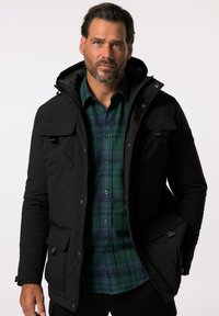 Schwarze isolierte Jacke mit Reißverschluss auf der Vorderseite, verstellbarer Kapuze und mehreren Taschen. Tragen über einem grün-blauen Karohemd.