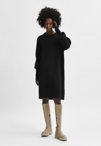 Liela izmēra melna vilnas kleita ar garām piedurknēm, saskaņota ar bēšiem ceļgala augstiem zābakiem ar sānu zipsi un masīvām zolēm.