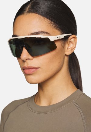 Donna con capelli scuri raccolti in una coda di cavallo, che indossa grandi occhiali sportivi neri con montatura bianca e una maglia girocollo verde oliva.