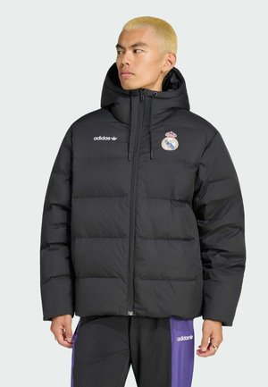 Hombre con cabello rubio corto que lleva una chaqueta acolchada negra de Adidas con el logo del Real Madrid y pantalones Adidas negros y morados, mirando hacia la izquierda.