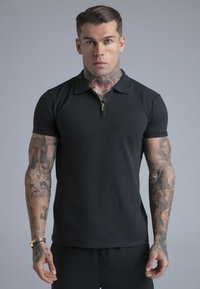 Hombre con tatuajes en el cuello y los brazos, vistiendo una camiseta tipo polo negra de manga corta con cremallera frontal, de pie frente a un fondo gris.