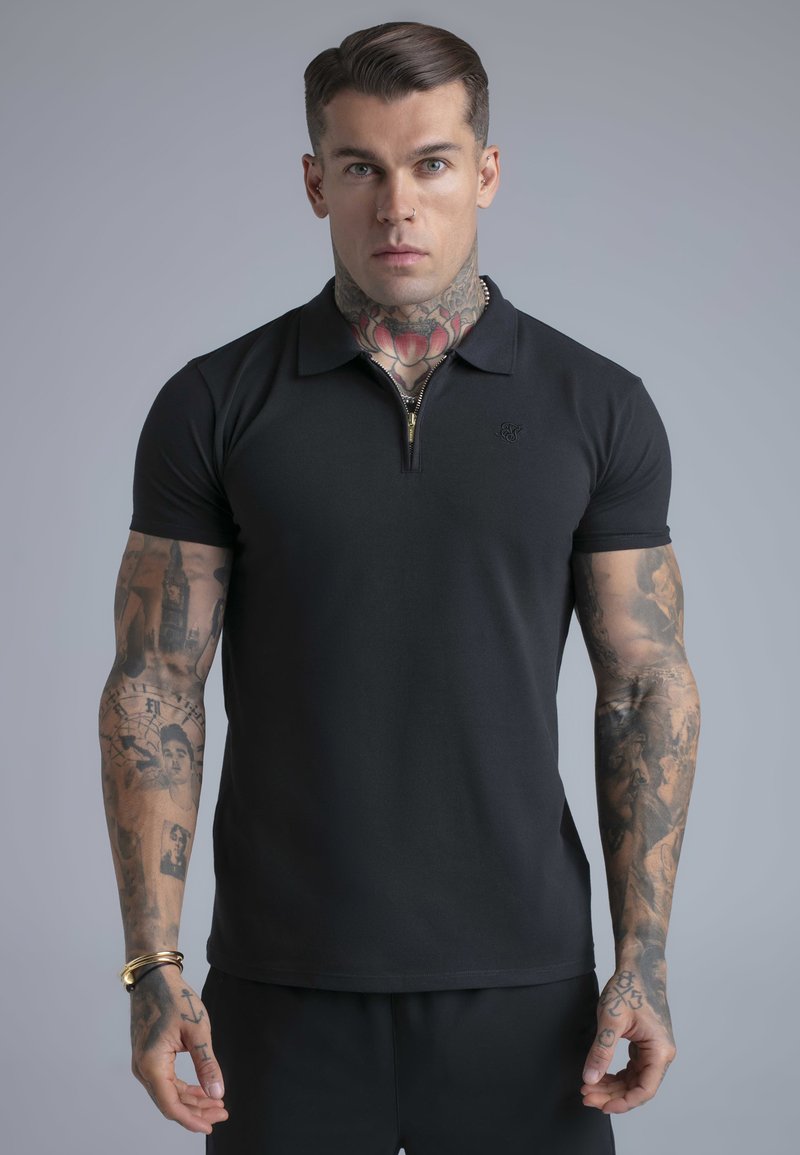 Hombre con tatuajes en el cuello y los brazos, vistiendo una camiseta tipo polo negra de manga corta con cremallera frontal, de pie frente a un fondo gris.