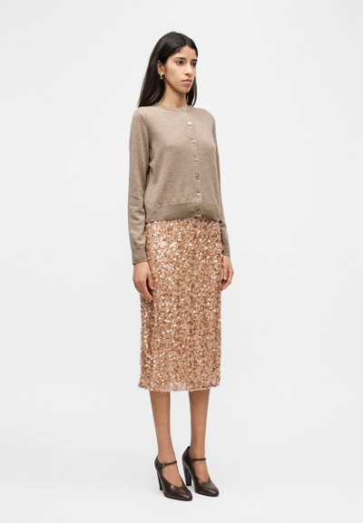 Μπεζ ζακέτα με στ ρογγυλή λαιμόκοψη και κουμπιά, σε συνδυασμό με φούστα από παγιέτες σε ροζ χρυσό χρώμα. Γυναίκα που φοράει καφέ παπούτσια με τακούνι.