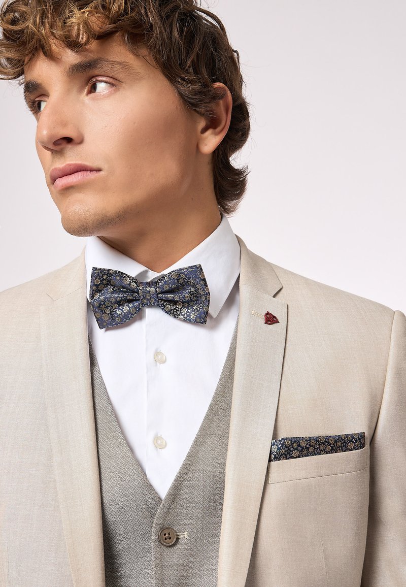 Giacca beige con camicia bianca e gilet grigio. Papillon con motivo floreale blu. Fazzoletto da taschino con un motivo simile. Distintivo accento rosso.