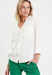 Chemise en lin blanche à manches longues avec un col à revers et deux poches poitrine, associée à un pantalon vert ; présente des boutons et une coupe décontractée.
