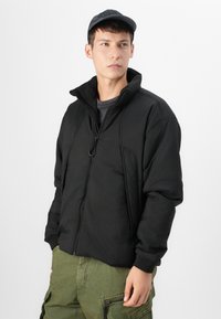 Veste zippée noire avec col haut et tissu texturé, dotée de poches latérales et de poignets côtelés, portée sur une chemise grise et un pantalon vert.