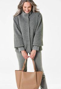 Grau strukturierter Pullover mit hohem Kragen, kombiniert mit hellgrauen Hosen. Hält eine hellbraune Tragetasche mit schlichter Gestaltung und vertikalen Taschendetails.