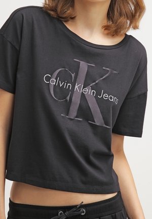 Vrouw draagt een zwart cropped Calvin Klein Jeans t-shirt met groot "CK" logo en zwarte broek met trekkoord, torso zichtbaar.