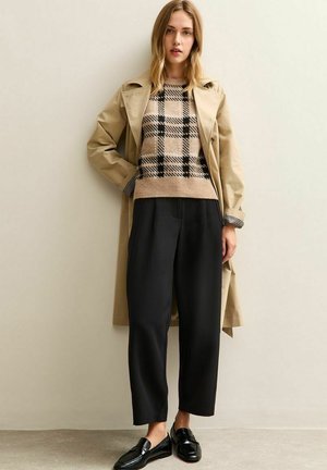 Beige trenchcoat over een bruin en zwart geblokt sweater, gecombineerd met zwarte wijde broek en zwarte loafers. Soepele texturen en een op maat gemaakte pasvorm.