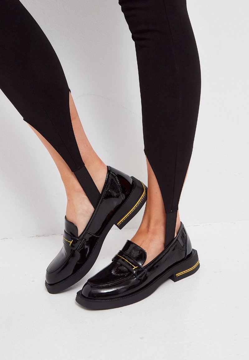 Mocassins en cuir verni noir avec un bout arrondi, une couture jaune et une semelle plate. Associés à des leggings noirs avec des découpes sur les côtés.