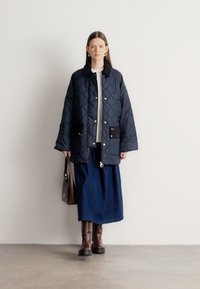 Gewatteerd marineblauw jack met zwarte accenten op de zakken, gedragen over een witte top en een lange marineblauwe rok, aangevuld met bruine kniehoge laarzen en een bruine tas.