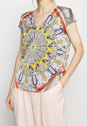Femme portant un haut à manches courtes avec un grand motif mandala complexe jaune, noir et orange et un pantalon beige clair.