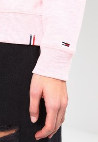 Lichtroze sweatshirt met geribbelde mouwen, voorzien van een klein geborduurd logo in rood, wit en blauw op de mouw. Hand rustend op zwarte stof.