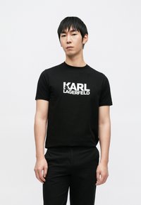 Fekete pamut póló fehér "KARL LAGERFELD" logónyomattal. Rövid ujjú, kerek nyakú, laza szabású. A modell fekete nadrággal viseli.