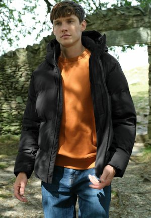 Sort pufferjakke med hætte, iført over en orange sweatshirt. Modellen bærer også blå jeans. Scenen foregår udendørs med naturligt lys.