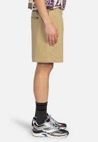 Pantalones cortos beige con un corte relajado, que cuentan con bolsillos laterales y una textura suave, combinados con zapatillas negras y blancas y calcetines oscuros.
