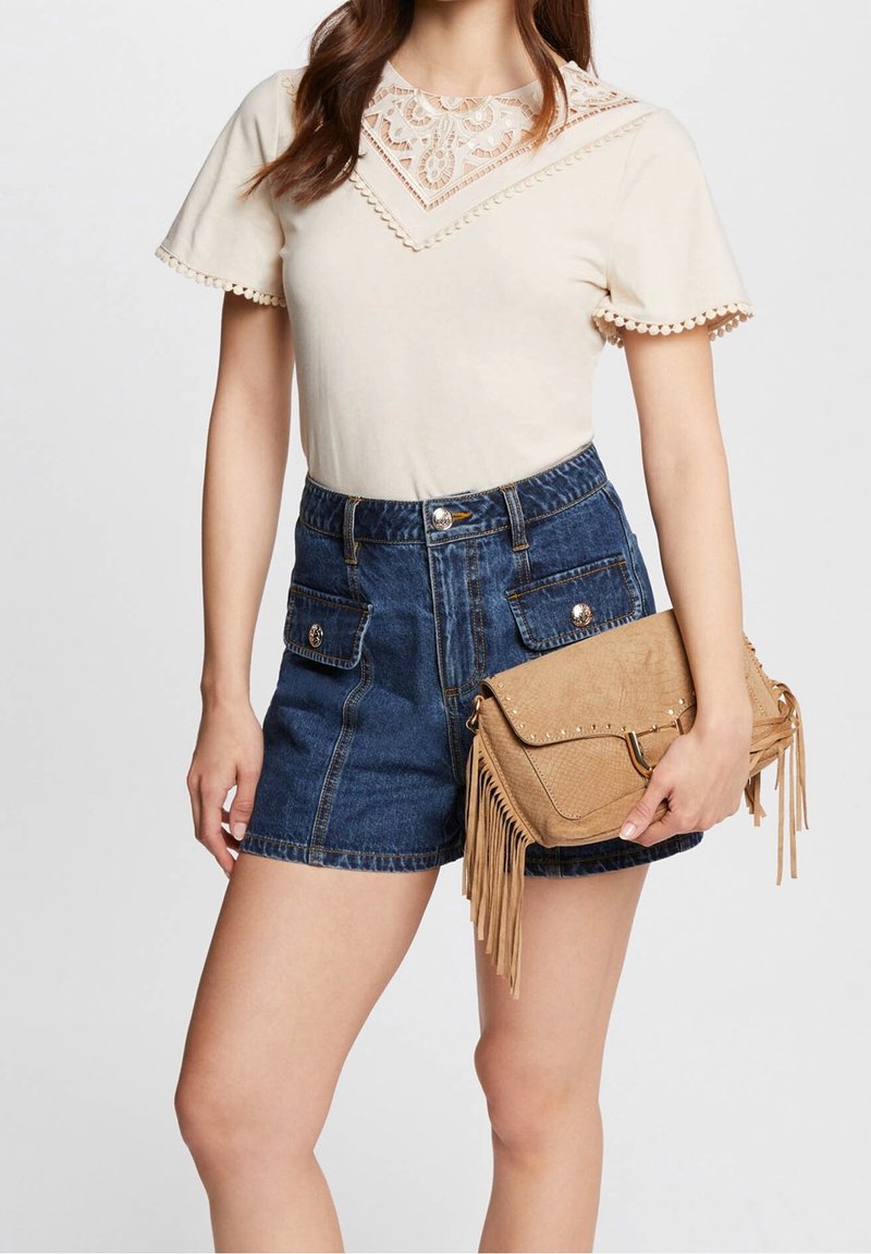 Femme portant un haut beige à manches courtes bordé de dentelle, un short en jean bleu taille haute avec poches, tenant un sac à main marron à franges avec des clous.
