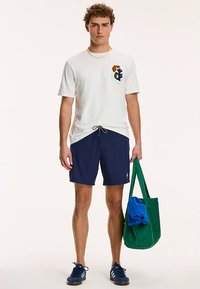 T-shirt en coton blanc avec logo abstrait, short de bain bleu marine, sac fourre-tout vert avec une serviette bleue à l'intérieur, et chaussures bleu marine. Modèle debout, en position verticale.