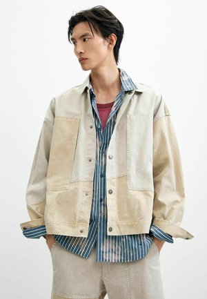 Ung mand med hænderne i lommerne iført beige patchworkjakke over blåstribet skjorte og vinrød t-shirt, kigger mod venstre.