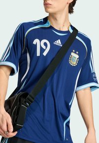 Giovane che indossa una maglia da calcio dell'Argentina blu con il numero 19, logo Adidas e tracolla nera a tracolla sul petto.