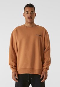 Felpa marrone realizzata in tessuto morbido, con scollo rotondo e maniche lunghe. Testo del logo nero sul petto sinistro. Vestibilità rilassata.